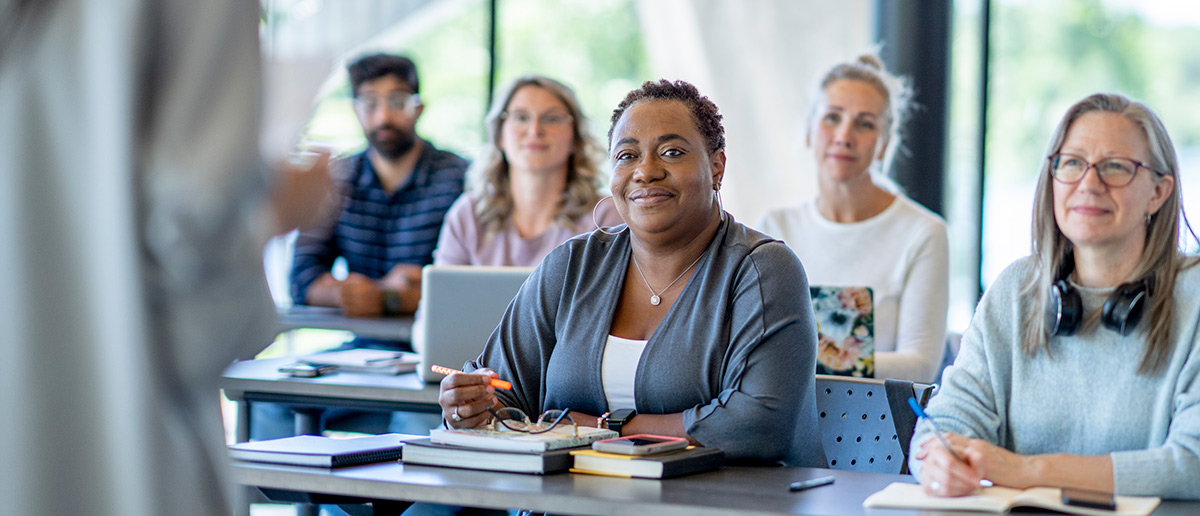 A large group of diverse mature students, sit together in rows as they listen attentively to their teacher explain the lesson. 