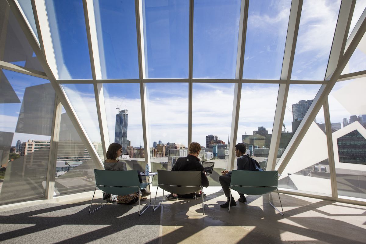 3 students sitting facing a large window showing a bright blue sky