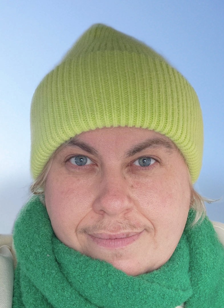 A white person with freckles and blue eyes. They are wearing a green toque and scarf outside.