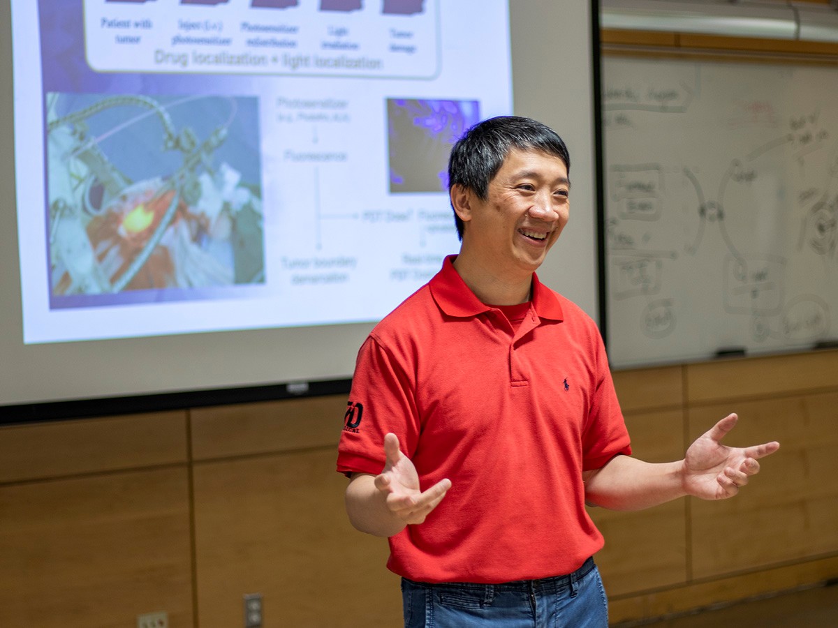 Professor Victor Yang smiles and stands in front of a whiteboard and projector screen in a classroom.