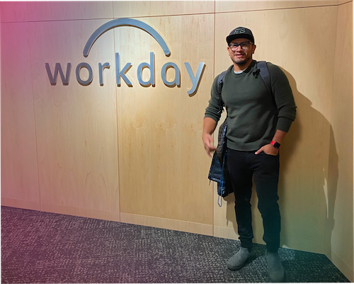 "A full-body photograph of a man, Fernando Toro, standing in an office environment. He is smiling and wearing a dark olive green crewneck sweater, black pants, grey boots, a black baseball cap, and glasses. He has a backpack on and a red watch on his left wrist. He is standing in front of a light-wood paneled wall featuring a large, silver 'workday' logo. The lighting is soft with a slight pink and teal hue cast from the sides."