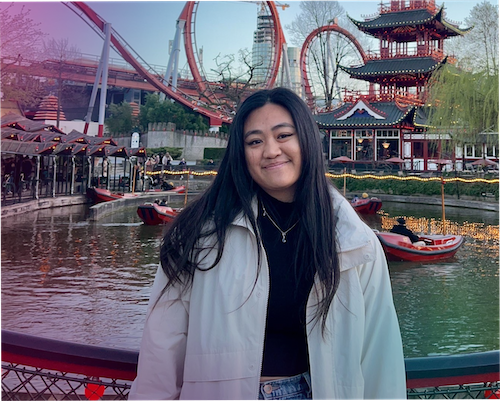 "A portrait of Natalie Cheung smiling in an amusement park setting. She has long, straight dark hair and is wearing a black top with a necklace, layered under an open off-white jacket. The background features an outdoor canal with small boats, roller coasters, and a traditional multi-tiered Asian pagoda building. The image has a transparent pink-to-purple colour gradient overlay on the upper left side."
