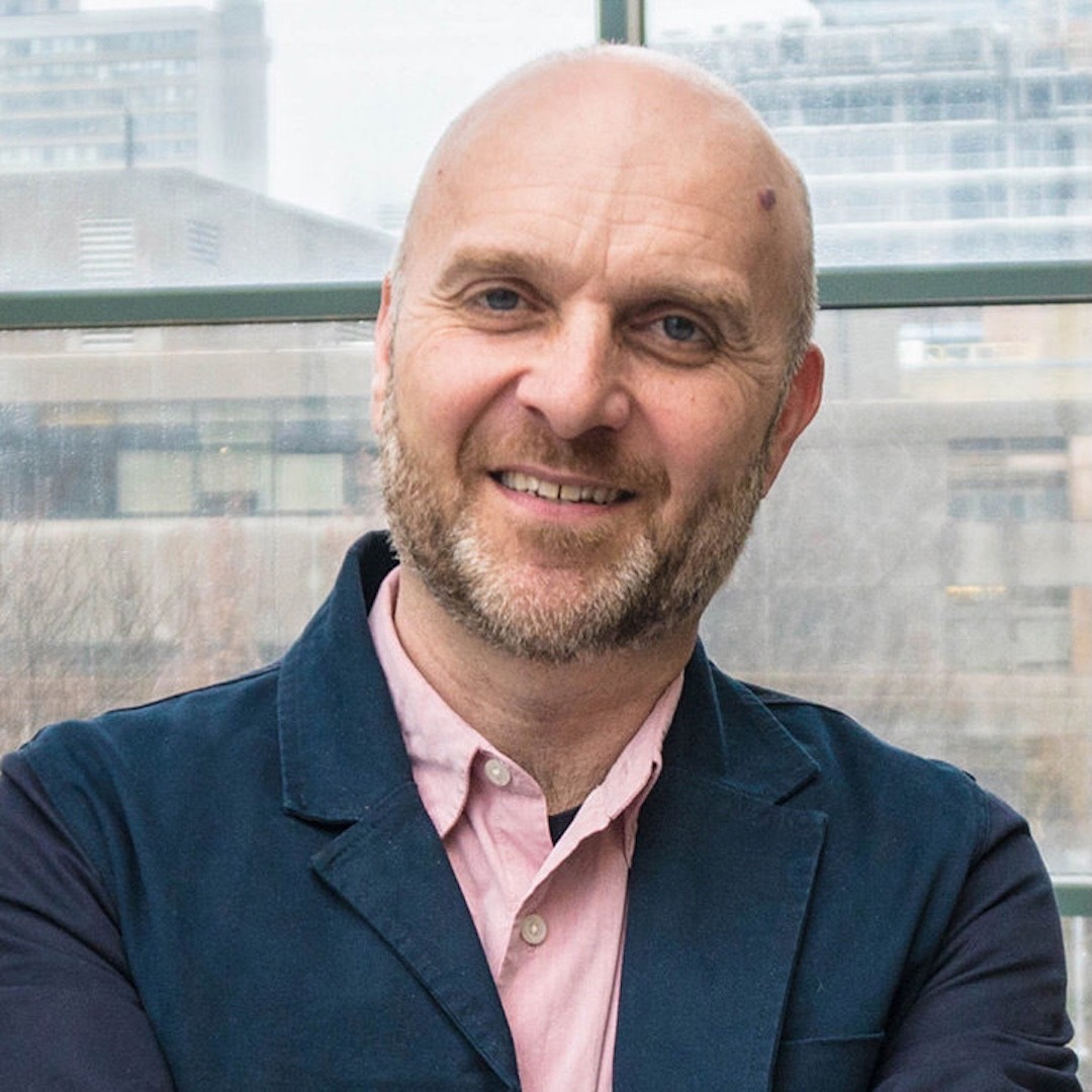 Close-up headshot of Greg Elmer. He is smiling and wearing a blue blazer with a pink button up underneath.