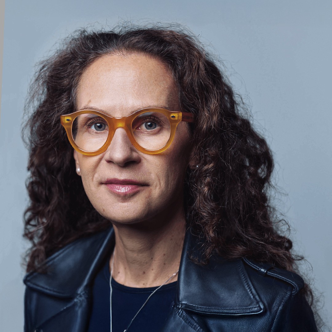Close-up headshot of Jessica Mudry. She is wearing orange-rimmed circular glasses with her brown hair down. She is wearing a leather jacket, a black shirt underneath. and a silver necklace.