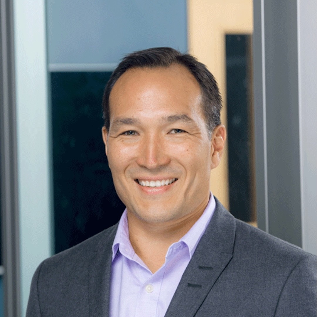 A close-up headshot of John Shiga in a gray blazer and light purple shirt. He smiles at the camera with short dark hair.