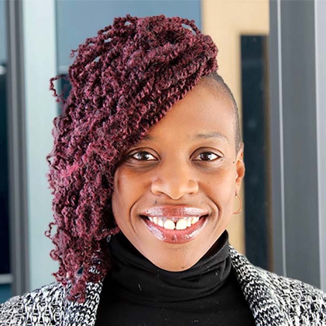 A close-up headshot of Kisha McPherson smiling person with curly, maroon-coloured hair swept to one side. She is wearing a black turtleneck and a grey patterned jacket.