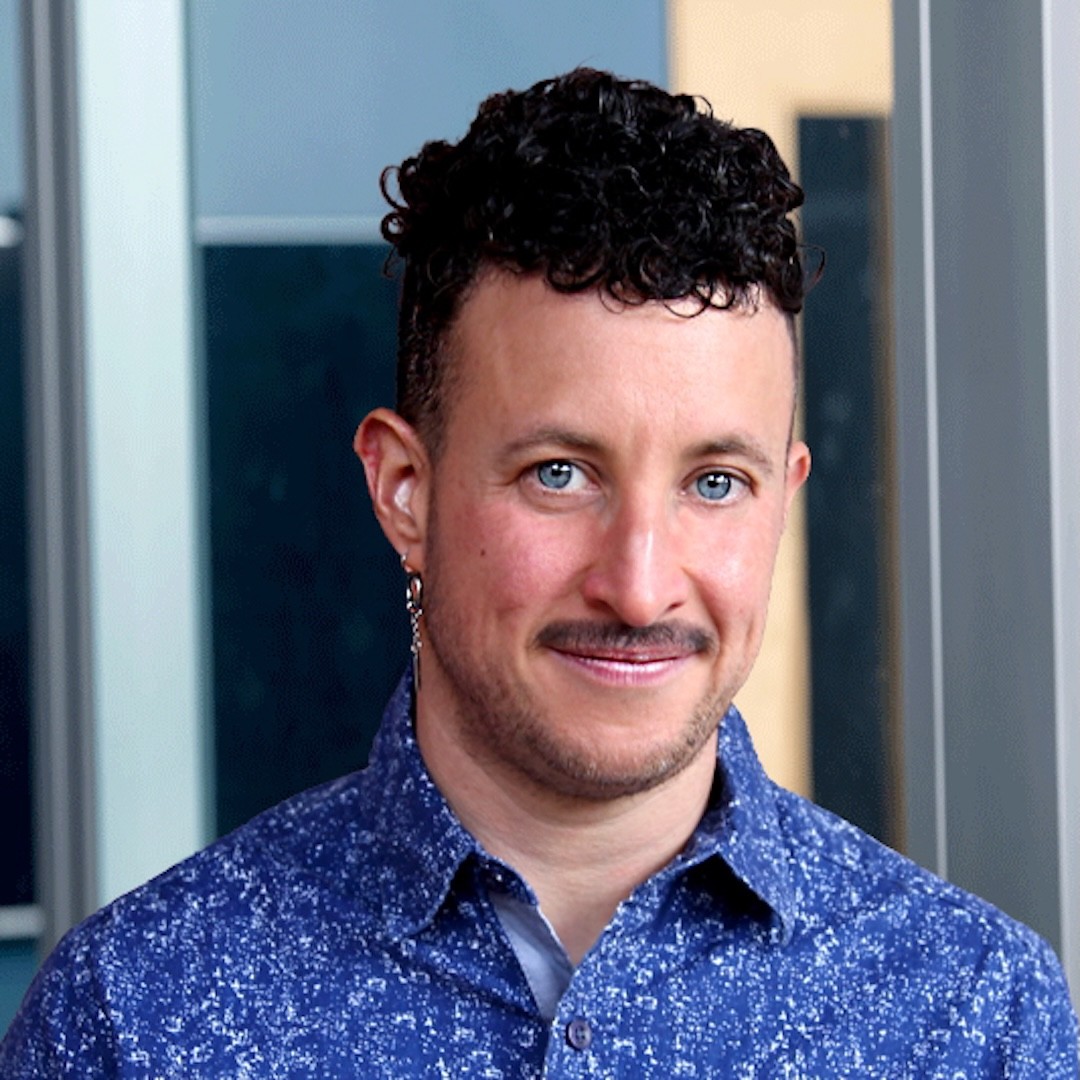 A close-up headshot of Marty Fink wearing a patterned blue button-up. He has short curly black hair, long silver earrings, and smiles slightly at the camera.