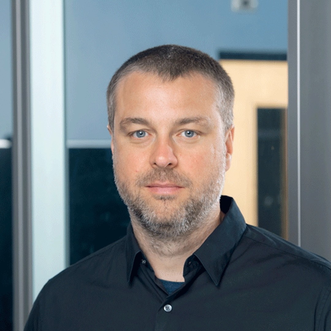 Close-up headshot of Matthew Tiessen. He wears a neutral expression and with a dark grey beard and moustache. He is wearing a dark blue button-up shirt. 
