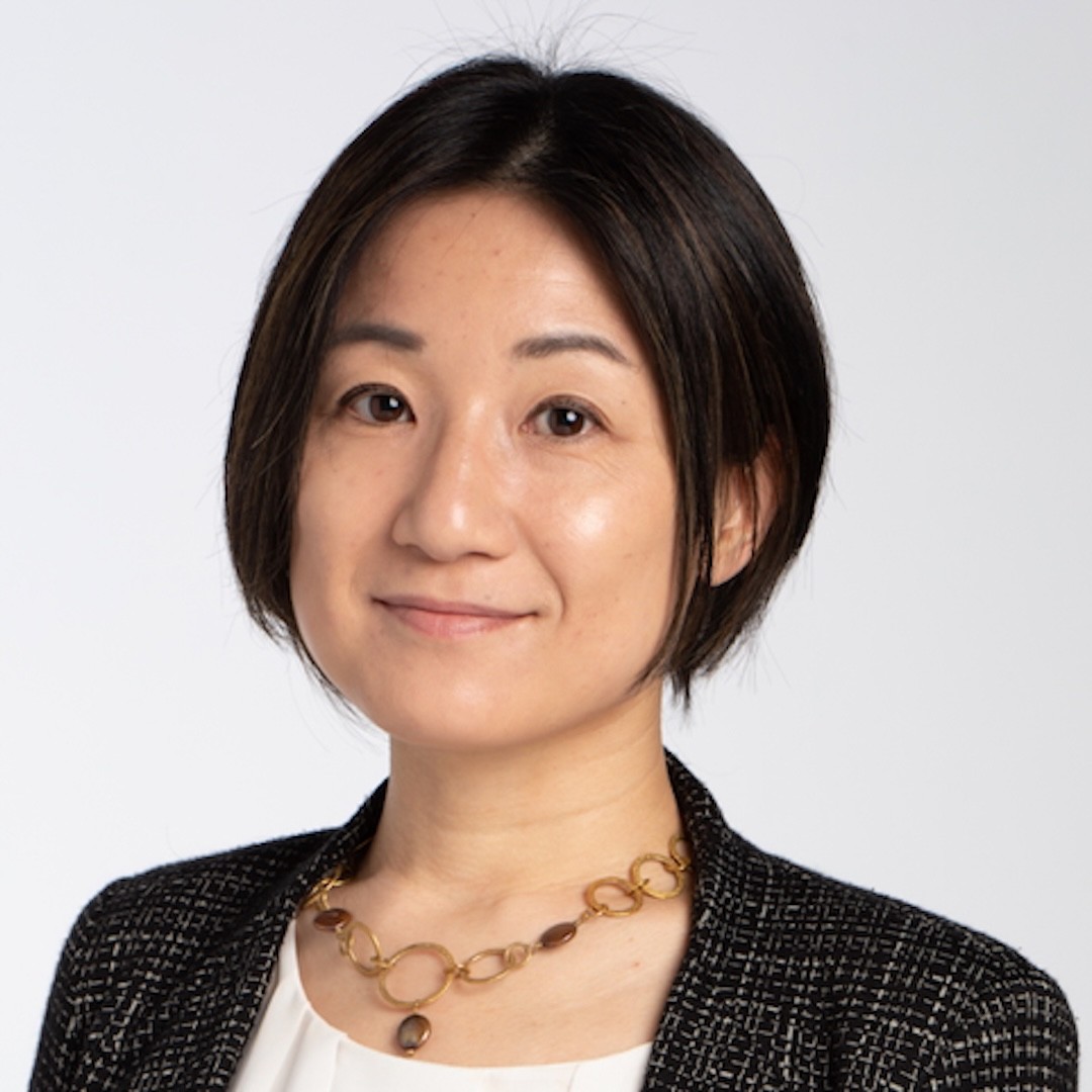 A close-up headshot of Yukari Seko with straight and short black hair. She wears a black textured blazer, a white blouse, and a gold chain necklace. She is smiling slightly against a plain light background.