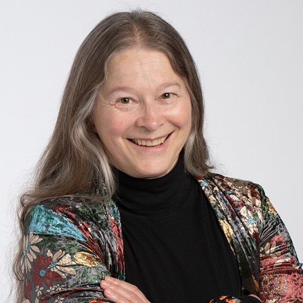 A close-up headshot of Catherine Jenkins with gray hair tied back and smiling. She wears a colourful scarf with a floral pattern, a white top, and a green jacket. 