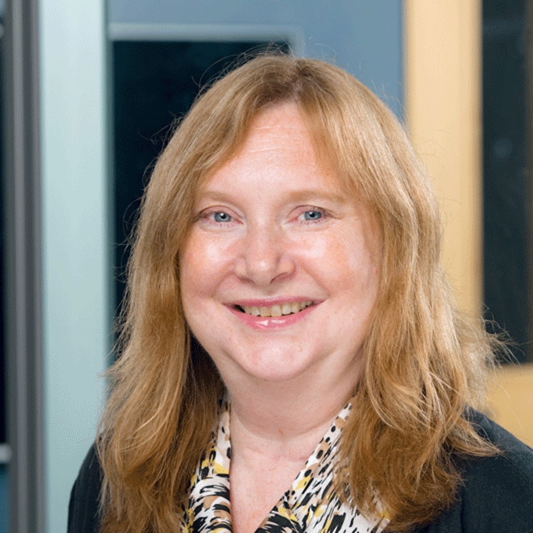 A close-up headshot of Linda Schofield with long, light brown hair smiling at the camera. She is wearing a black jacket over a patterned blouse.