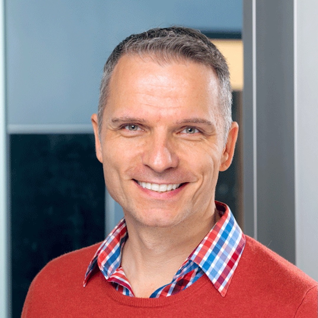 A close-up headshot of Robert Bajko smiling with short grey hair wearing a red sweater over a blue and red plaid shirt.