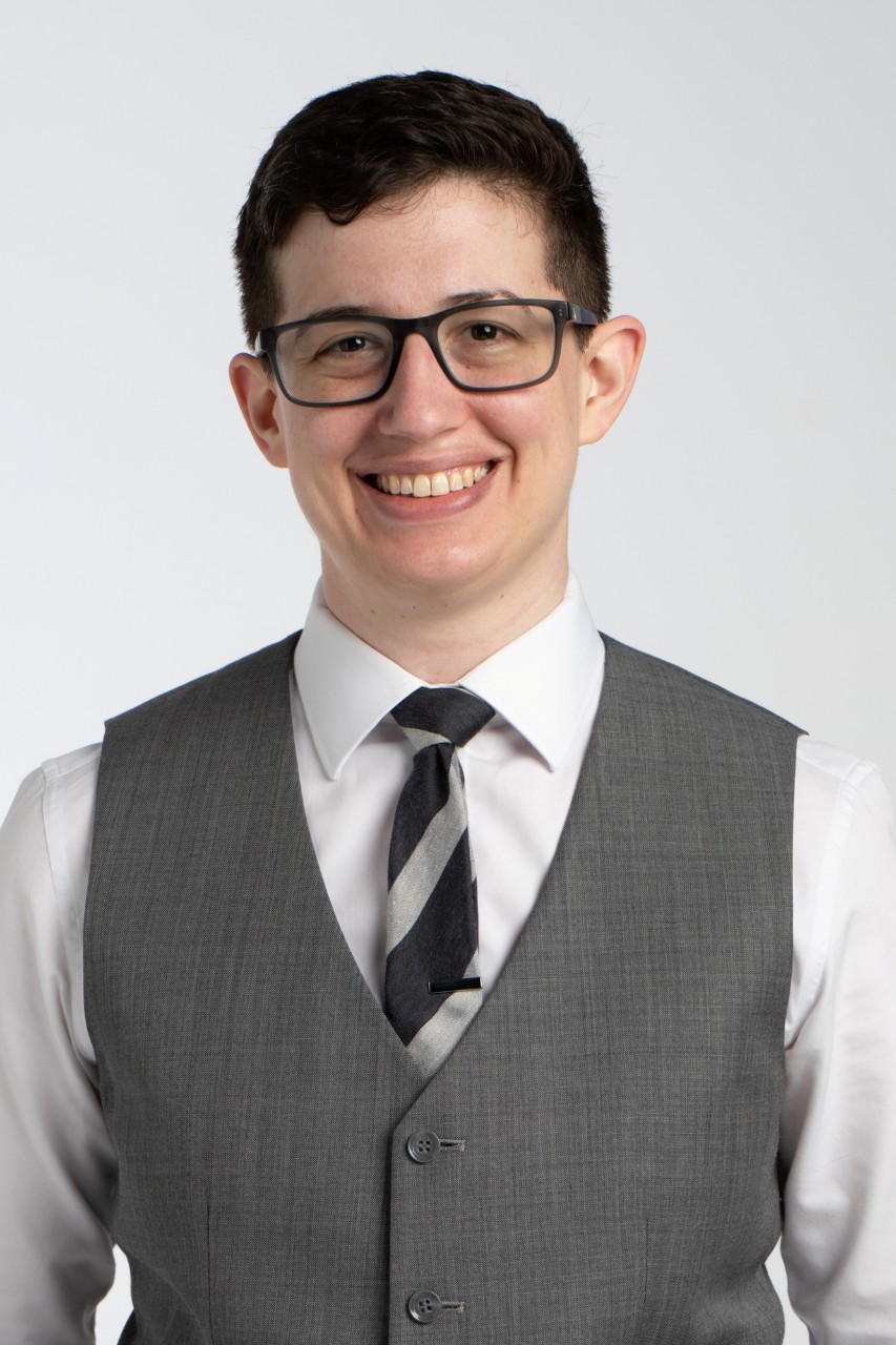 A close-up headshot of Sarah Kriger. She has short brown hair, glasses, and is wearing a white dress shirt, a black and silver tie, and a grey vest. 
