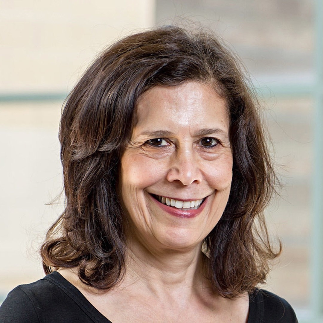 A close-up headshot of Sheila Rosenberg smiling with shoulder-length dark brown hair. She is wearing a black top.