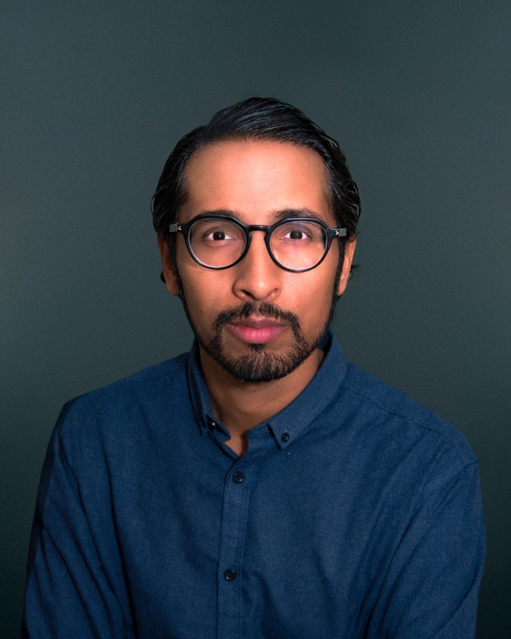 Harry looks at the camera. He has a striped shirt under a black blazer. He has dark hair and a goatee.