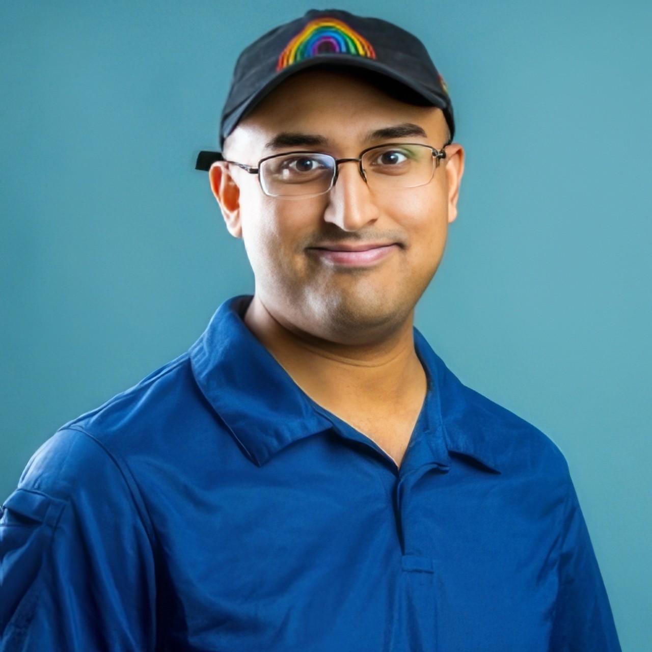 Close-up headshot of AJ Cordeiro smiling broadly. He is wearing a blue T-shirt with a black hat against, against a light blue background.