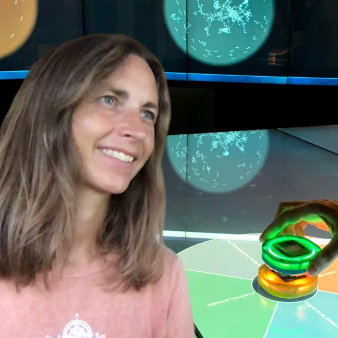Close-up headshot of Ali Mazalek smiling. She has shoulder-length hair parted in the middle and is wearing a light pink top. A colorful interactive display is in the background.