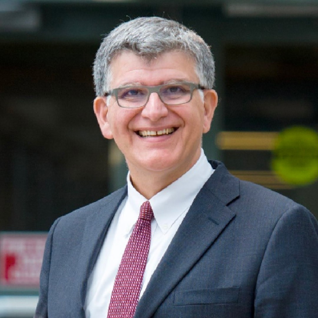 Close-up headshot of Charles Falzon smiling. He has short grey hair, glasses, and is wearing a dark suit with a white shirt and red tie.