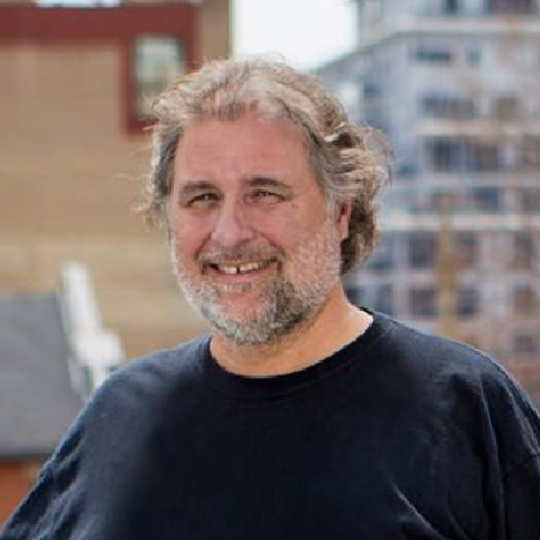Close-up headshot of Charles Zamaria smiling. He has curly grey hair and a beard and is wearing a black crew-neck shirt. Urban buildings are visible in the background.