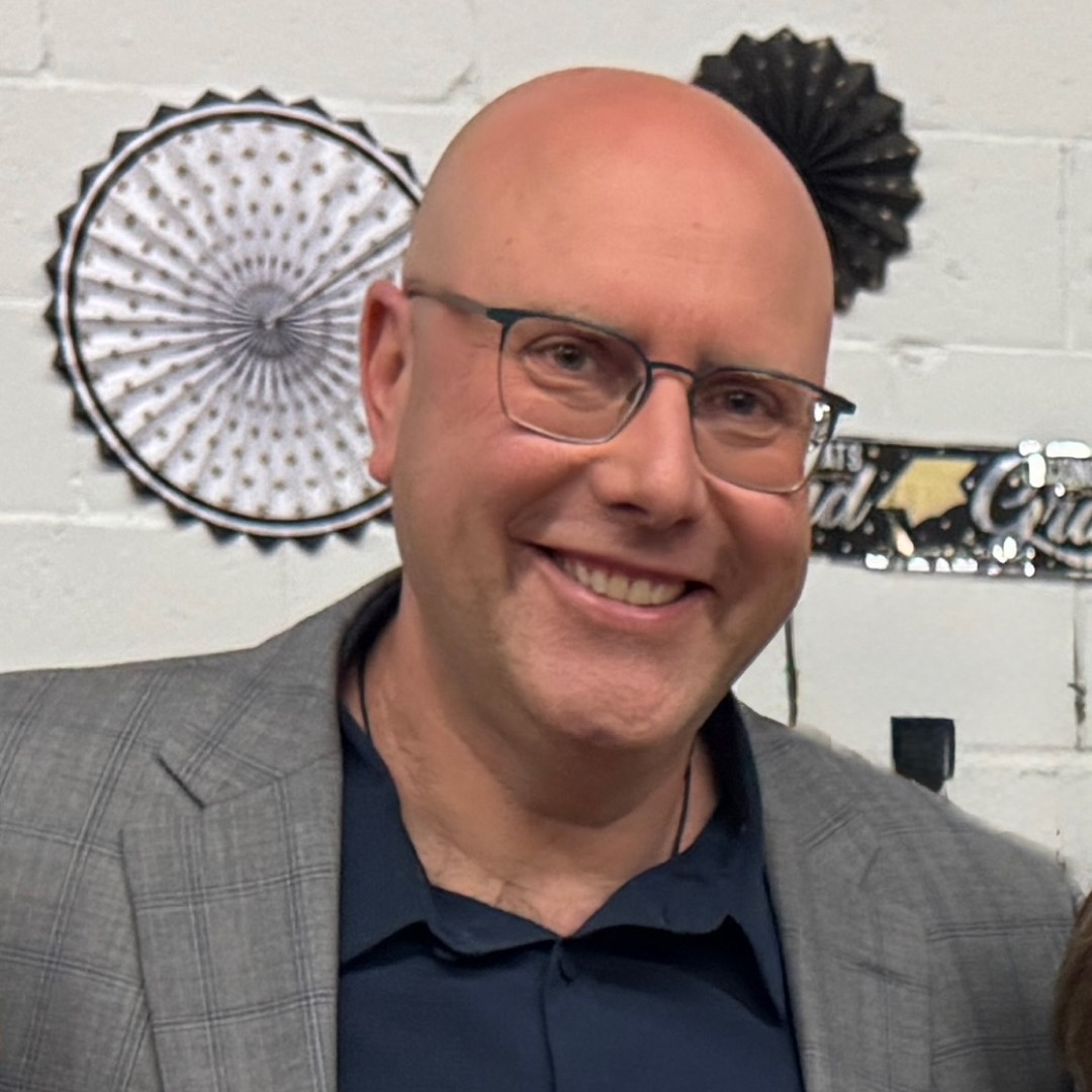 Close-up headshot of Dan Berlin smiling. He is bald, wearing glasses, and dressed in a navy shirt with a grey plaid blazer.