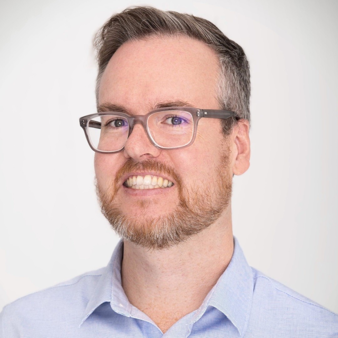 Close-up headshot of Finlay Braithwaite smiling. He has short hair, glasses, and a beard, and is wearing a light blue collared shirt.