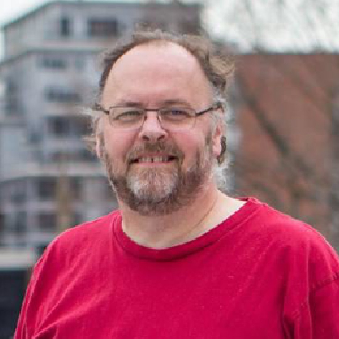 Close-up headshot of Henry Warwick smiling. He has glasses, a beard, and is wearing a bright red shirt.