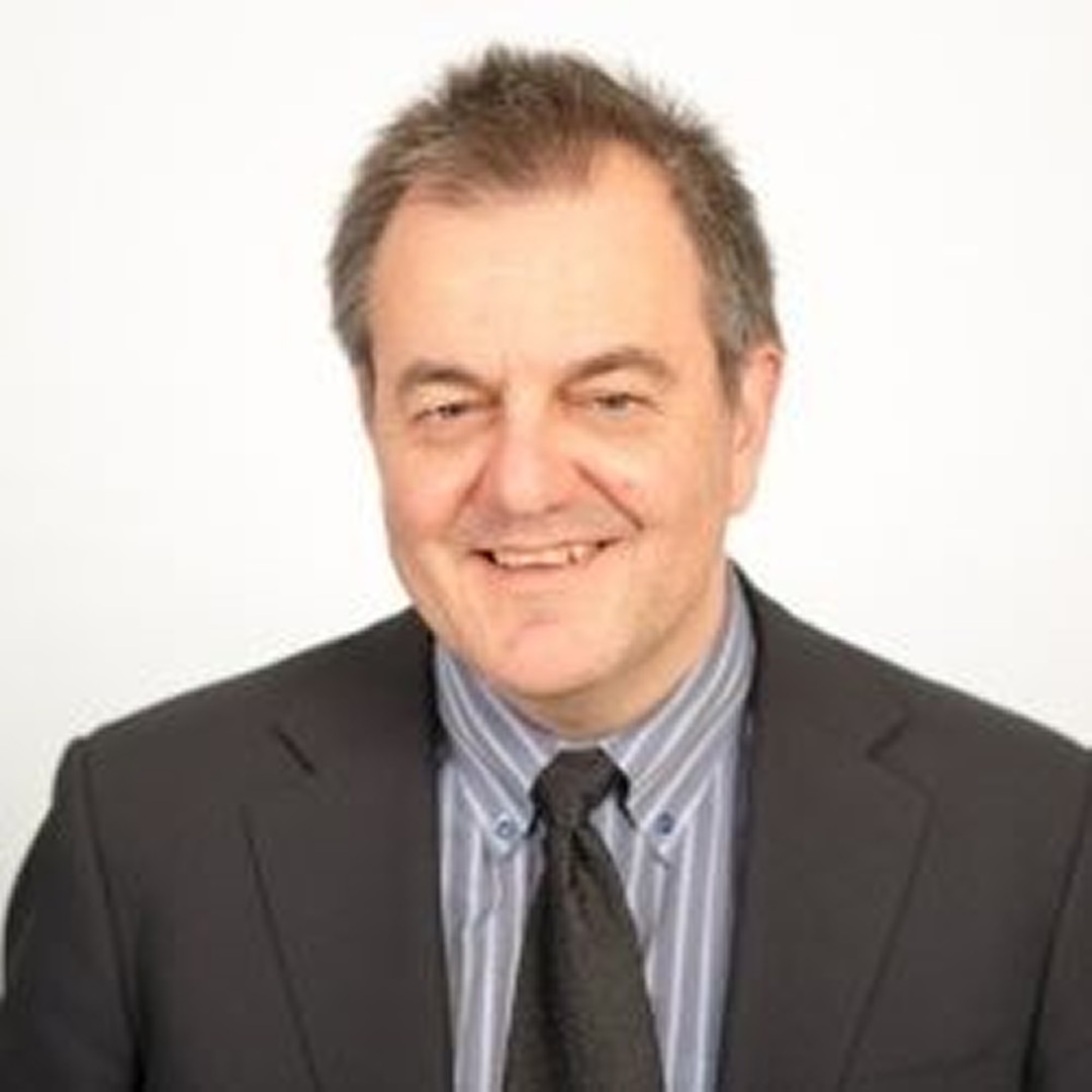 Close-up headshot of Joseph Recupero with a neutral expression. He has a beard and short dark hair and is wearing a green jacket over a pink collared shirt. A tree trunk is in the background.