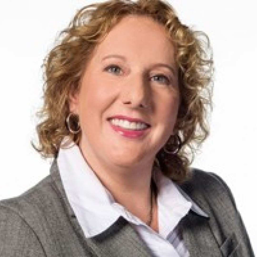 Close-up headshot of Karen Sebesta smiling. She has curly blonde hair and is wearing a grey blazer over a white collared shirt with hoop earrings.