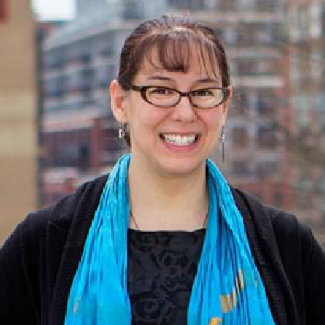 Close-up headshot of Kim Chow-Morris smiling. She is wearing glasses, earrings, a black top, a black cardigan, and a bright blue scarf.