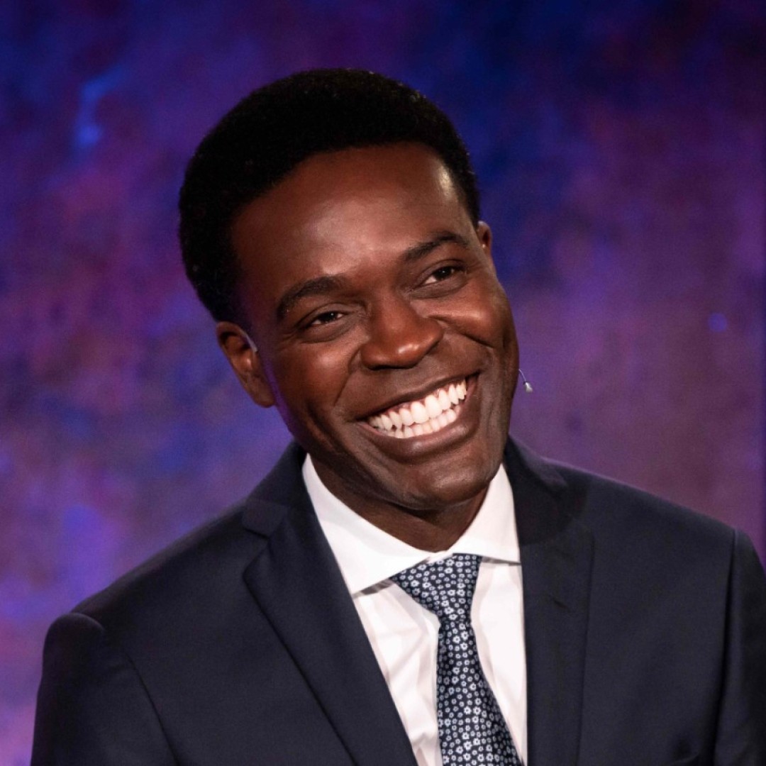 Close-up headshot of Kristopher Alexander smiling broadly. He is wearing a dark suit with a white shirt and patterned tie, against a purple background.