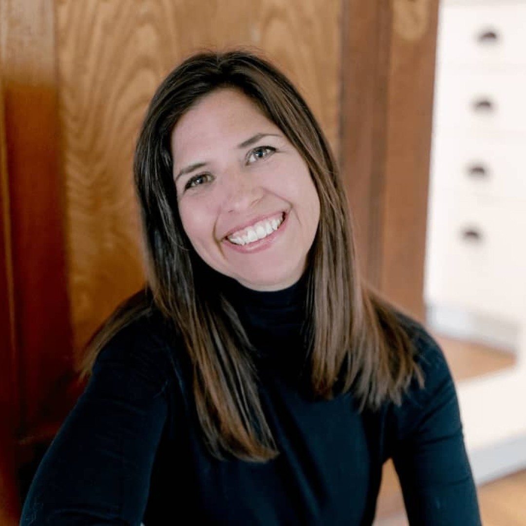 Close-up headshot of Laurie Petrou smiling. She has straight dark brown hair and is wearing a black long-sleeve turtleneck.