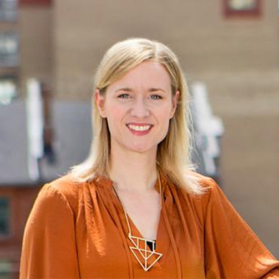 Close-up headshot of Lori Beckstead smiling. She has blonde hair parted to the side and is wearing an orange blouse with a geometric necklace.