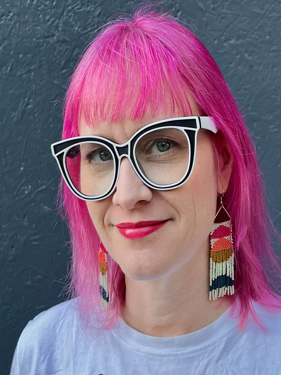 Close-up headshot of Lori Beckstead smiling. She has blonde hair parted to the side and is wearing an orange blouse with a geometric necklace.