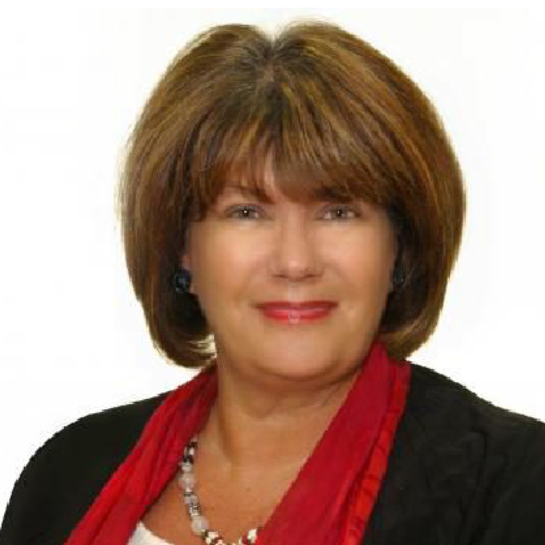 Close-up headshot of Marion Coomey smiling. She has a short layered hairstyle and is wearing a black jacket with a red scarf and beaded necklace.