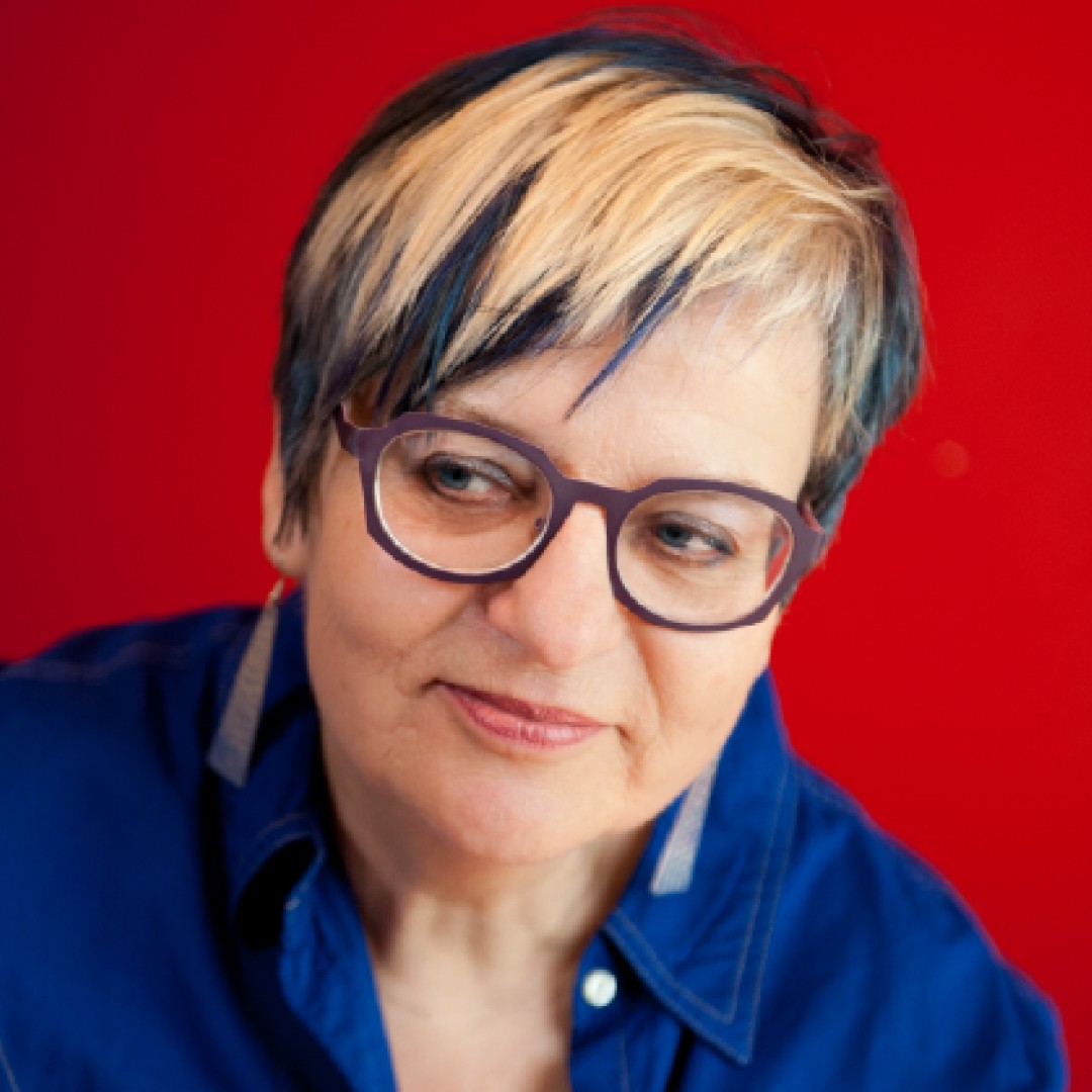 Close-up headshot of Marusya Bociurkiw looking slightly to the side with a soft smile. She has short hair with blonde highlights, glasses, and is wearing a blue collared shirt.