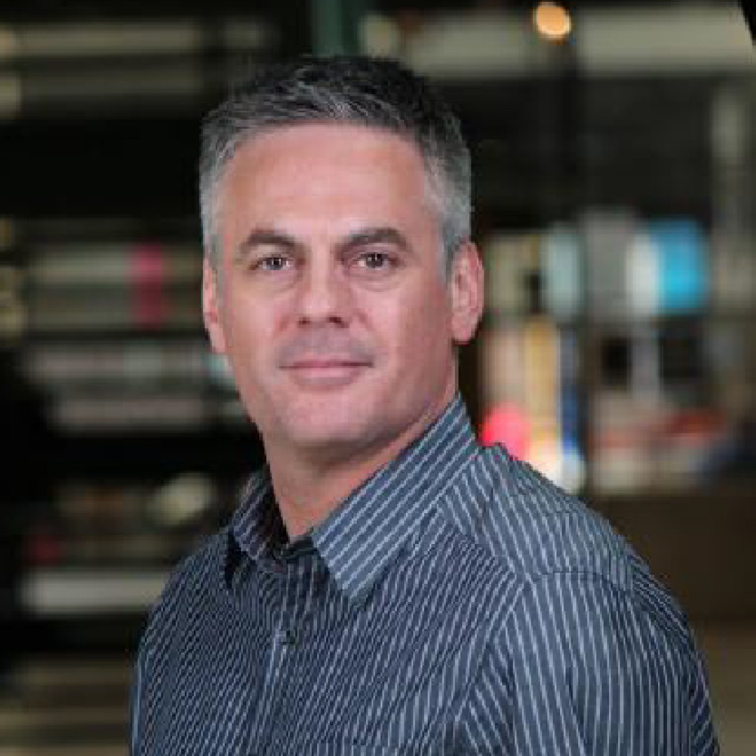 Close-up headshot of Michael Coutanche with a neutral expression. He has short grey hair and is wearing a striped button-up shirt.