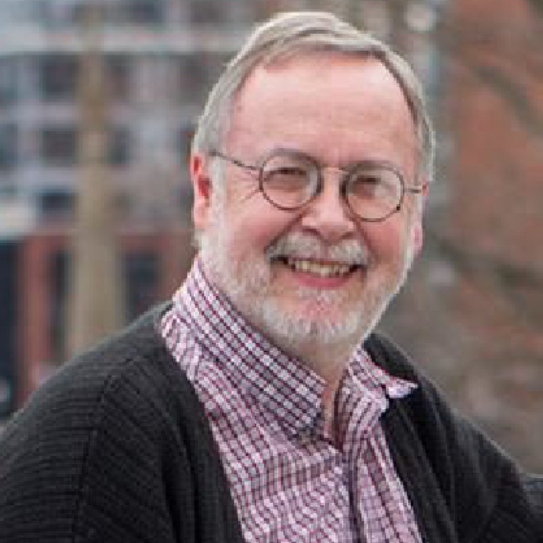 Close-up headshot of Michael Murphy smiling. He has grey hair, a beard, and glasses, and is wearing a checkered shirt with a dark cardigan.