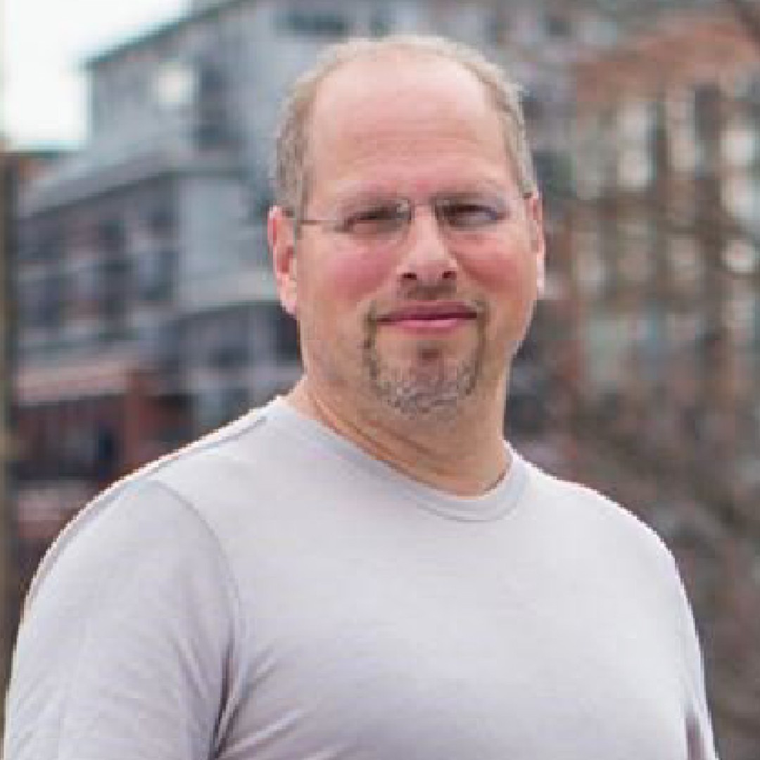 Close-up headshot of Richard Grunberg with a neutral expression. He is wearing glasses and a light grey T-shirt.