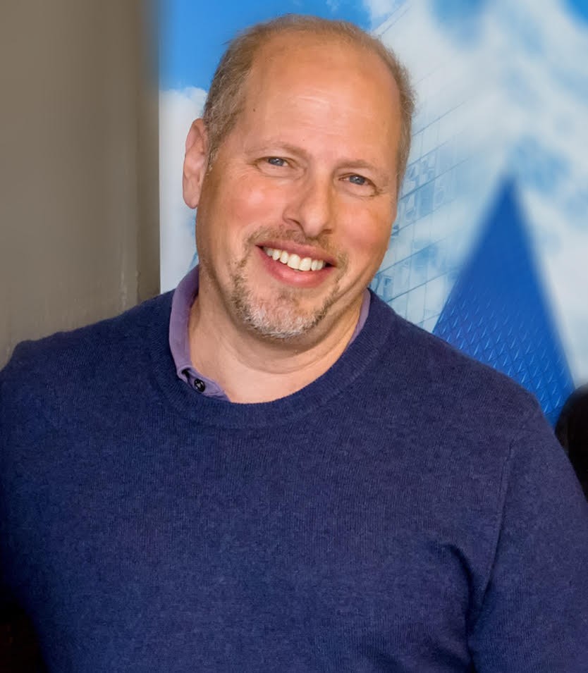 Close-up headshot of Richard Grunberg with a neutral expression. He is wearing glasses and a light grey T-shirt.