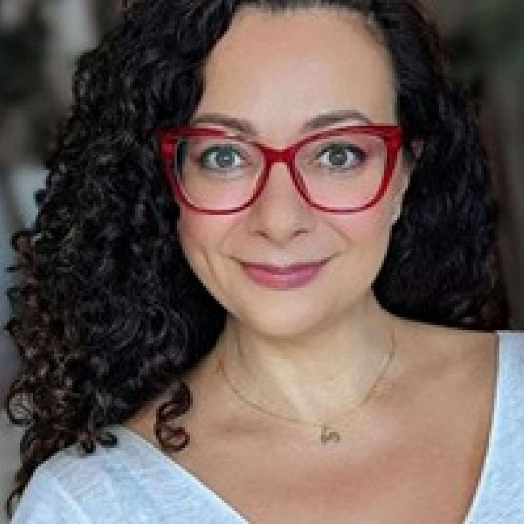Close-up headshot of Tatyana Terzopoulos smiling. She has curly hair, is wearing red glasses, and a white top with a necklace.