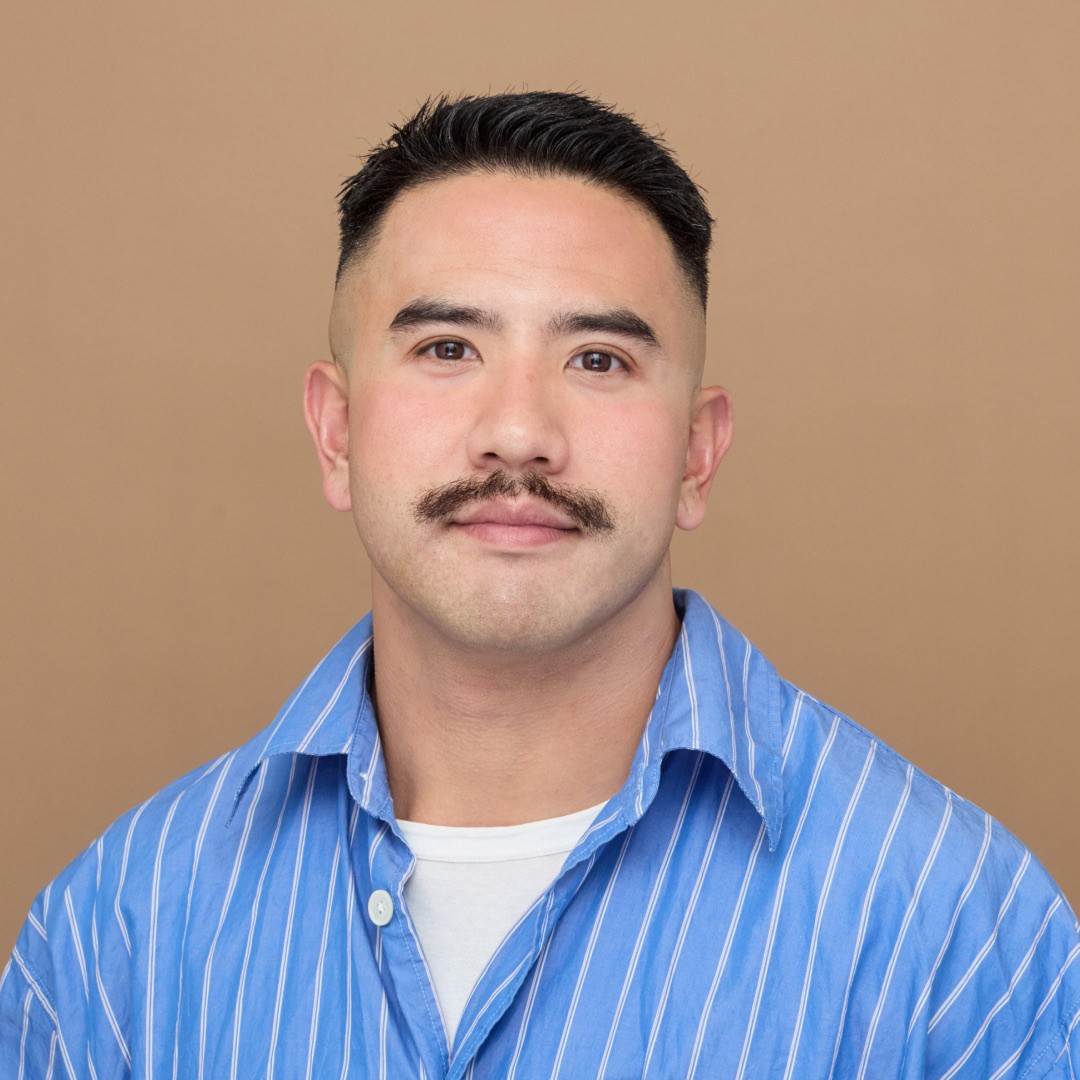 A close-up headshot of Tommy Ting. He has short black hair, a trimmed mustache, and is wearing a blue and white striped button-up shirt over a white undershirt. He is facing the camera with a neutral expression, against a plain tan background.